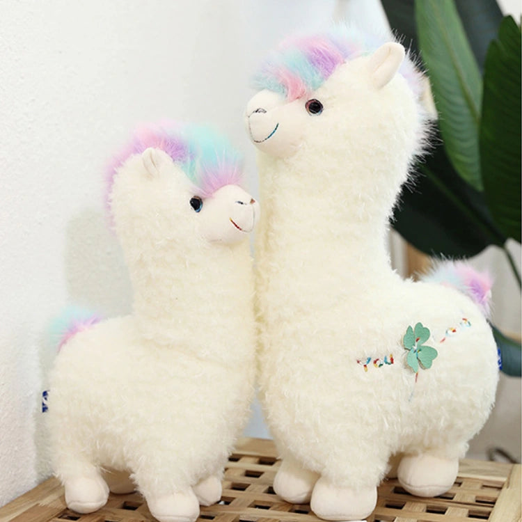Two ZoDoll Cute Alpaca Plush Toy - Soft Stuffed Alpaca Plush Dolls with pastel rainbow manes and tails stand side by side on a wooden surface, with a plant in the background.