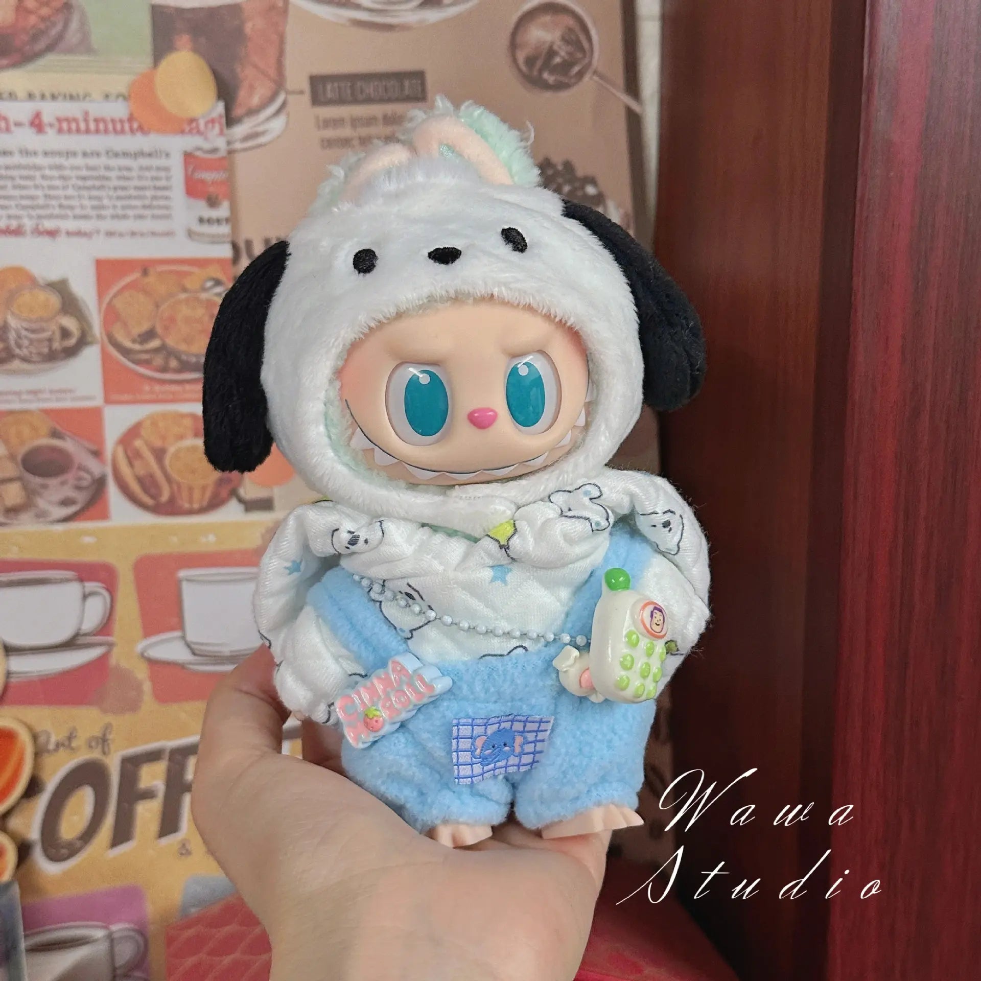 A hand holds a ZoDoll Labubu Sanrio Doll in a Hello Kitty Style Outfit—white dog-eared hood and blue shorts—from the Labubu Sanrio Doll Clothes Set, against a cafe background. The doll has big blue eyes and holds a toy phone.