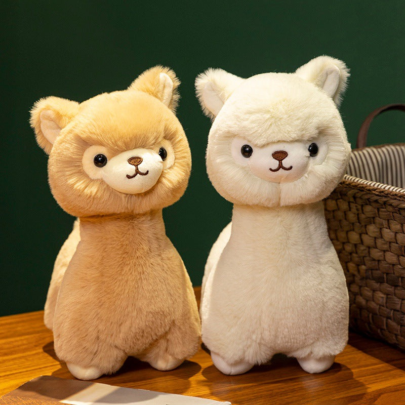 Two ZoDoll Cute Alpaca Plush Dolls—one light brown, one white—stand upright on a wooden surface by a woven basket, set against a dark green background.