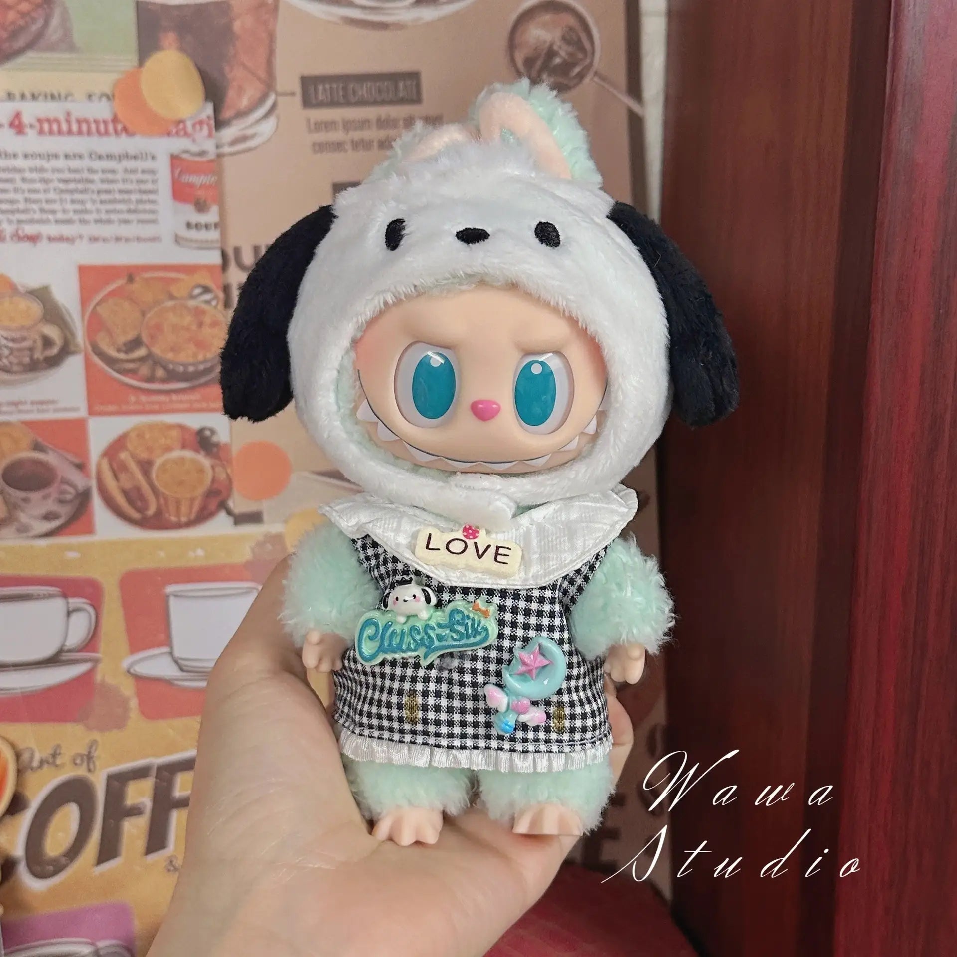 A person holds a ZoDoll Labubu Sanrio Doll Clothes Set – Hello Kitty Style Outfit Collection, featuring a mint green dog-eared hood, big blue eyes, and a patchwork checkered dress. A cafe-themed wall is in the background.