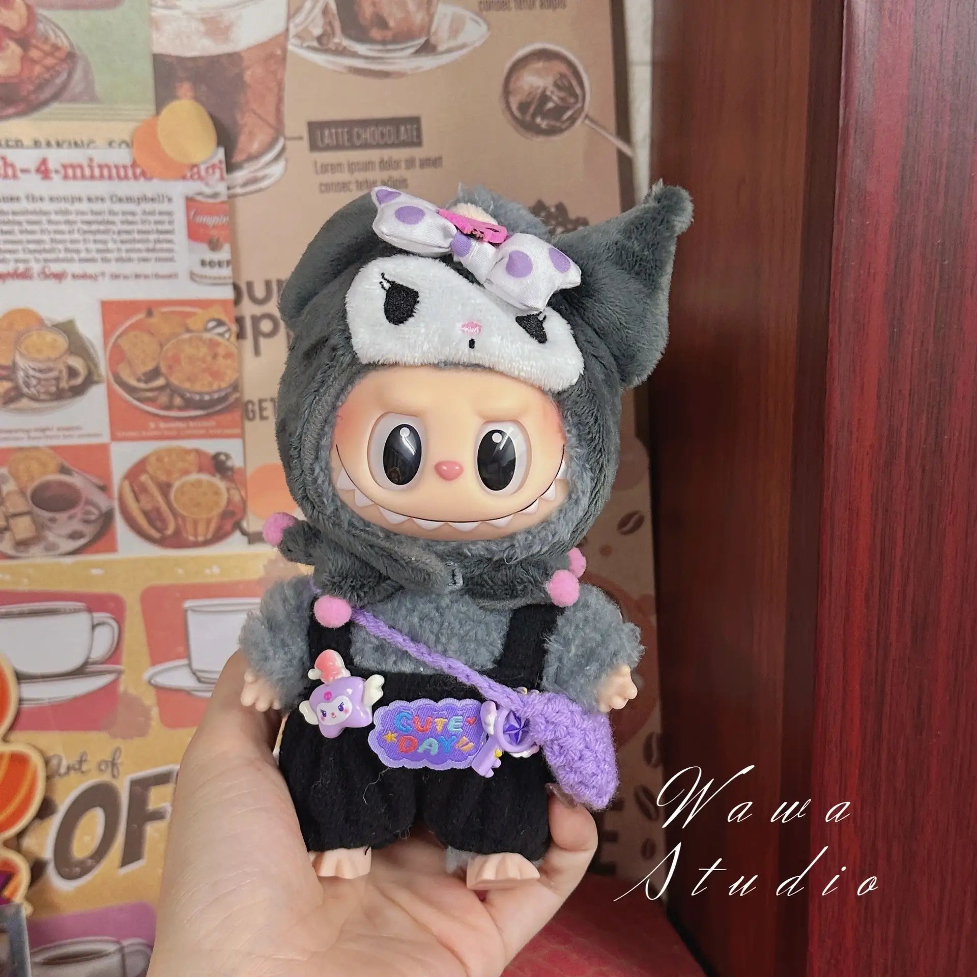 A hand holds the ZoDoll Labubu Sanrio Doll Clothes Set – Hello Kitty Style Outfit Collection, featuring a kawaii gray and black animal hoodie, bow, and purple bag, with a food-themed decor wall in the background.
