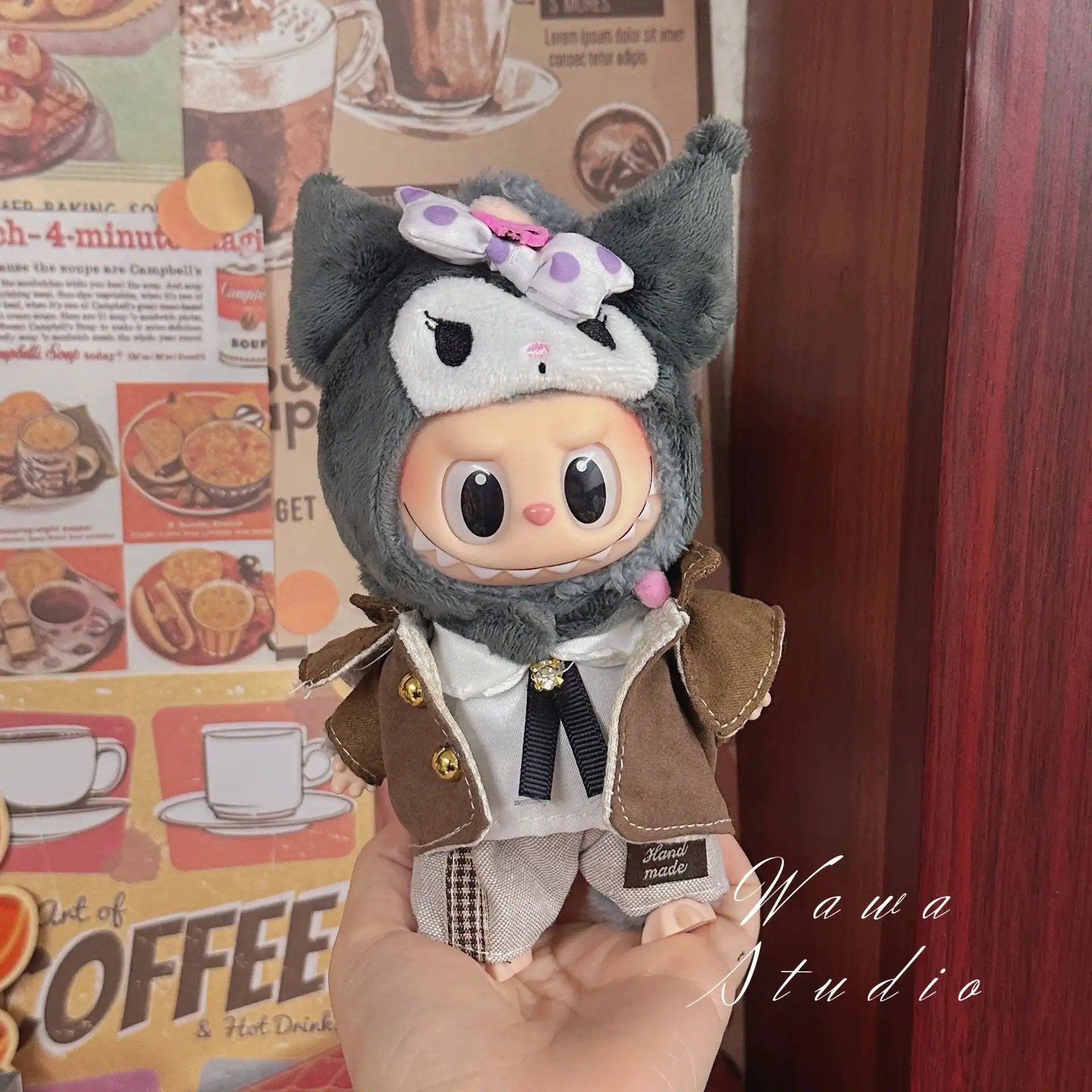A hand holds a ZoDoll Labubu Sanrio Doll dressed in the Hello Kitty Style Outfit Collection, featuring a brown coat and gray animal-themed hood with ears and a bow, posed before coffee-themed wall posters.