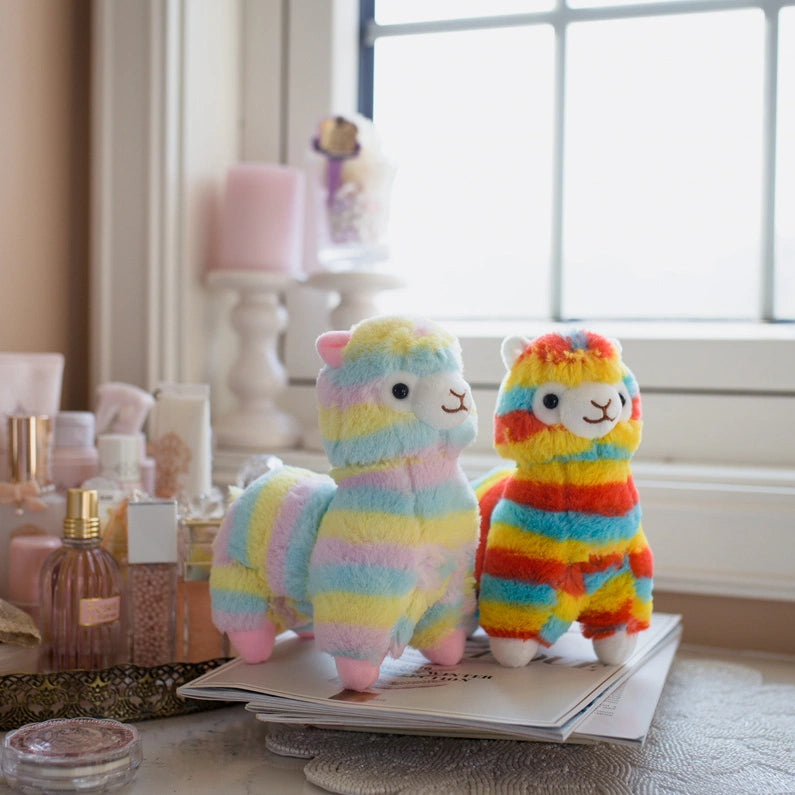 The ZoDoll Rainbow Alpaca Plush Toy - Cute Stuffed Alpaca Plush Doll adds a pop of color on a stack of magazines by a window, surrounded by perfume bottles and decorative items.