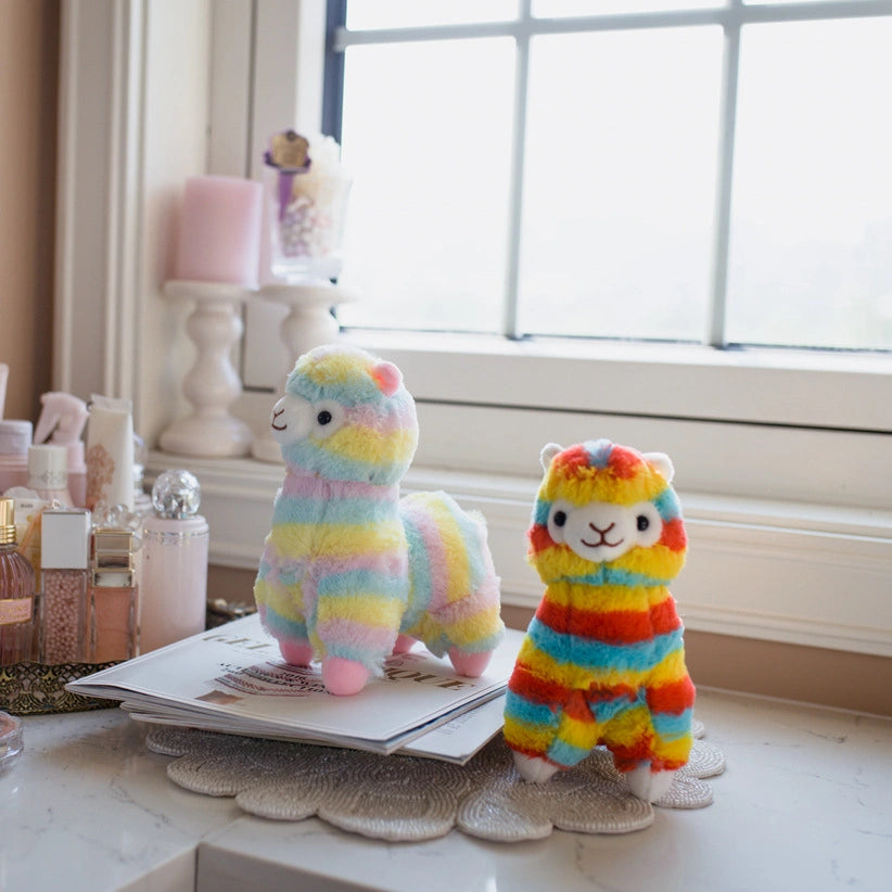 Two ZoDoll Rainbow Alpaca Plush Toys stand on a desk near a window, surrounded by decorative items and cosmetics.