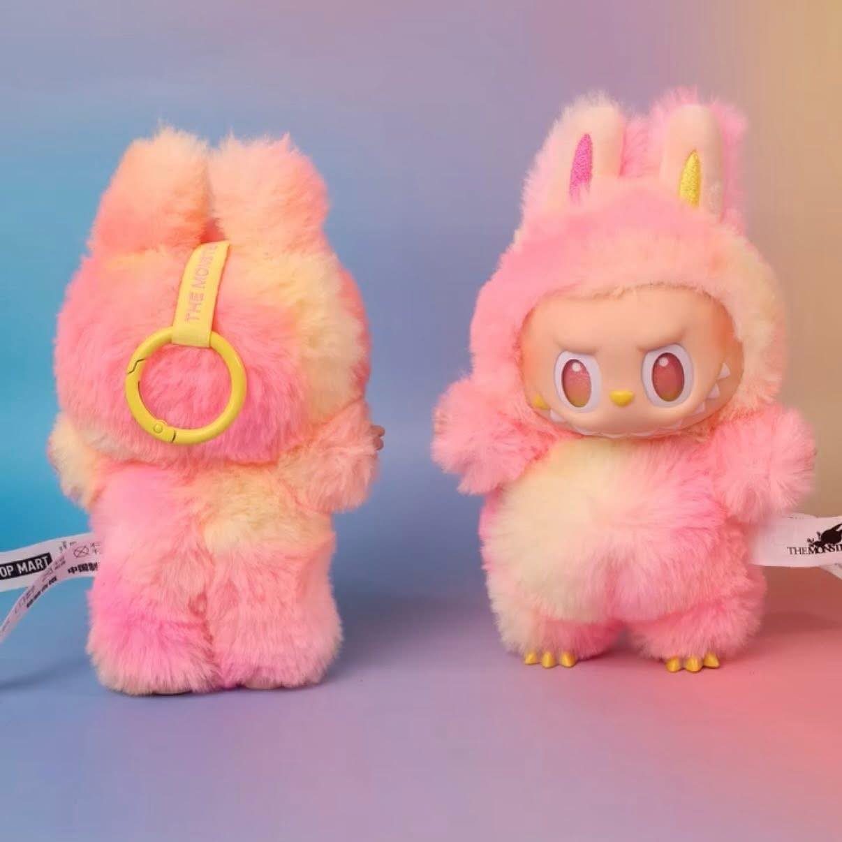 Two POP MART Labubu “Loyalty” Plush Keychains from The Monsters “Big Into Energy” Tie‑Dye Bunny Bag Charm series, featuring pink and yellow bunny fur, ears, and clawed feet, are shown front and back on a pastel background.