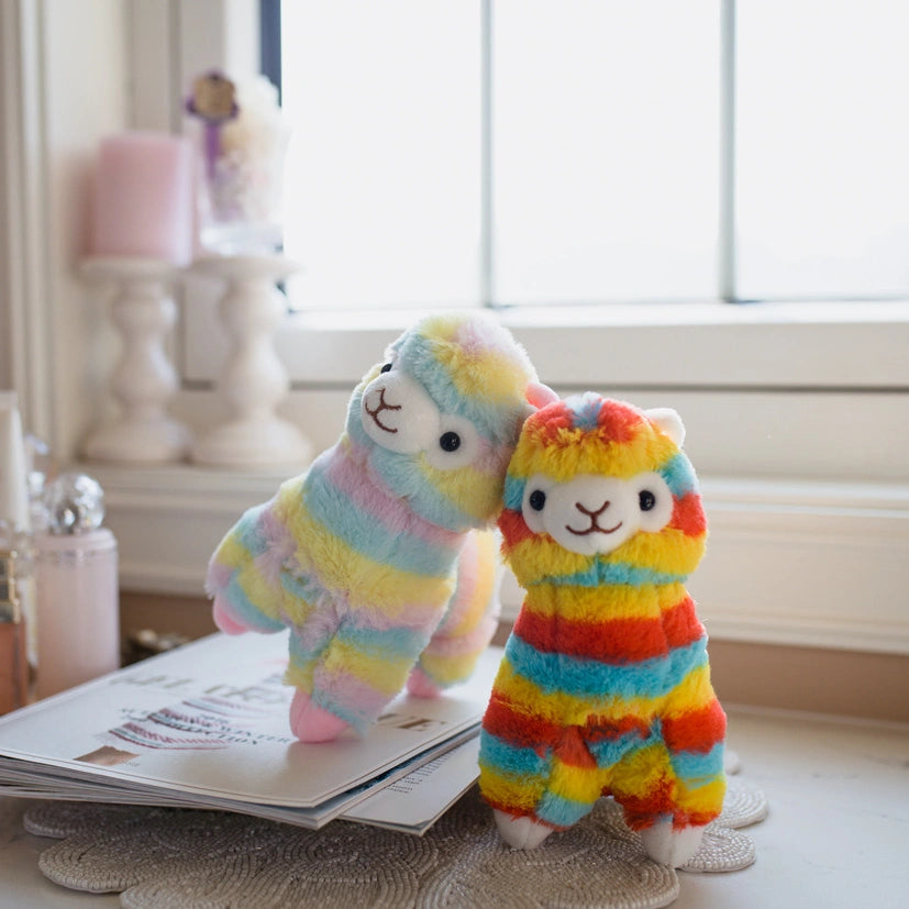 Two ZoDoll Rainbow Alpaca Plush Toys, featuring a cute rainbow and a pastel kawaii stuffed alpaca doll, stand on a magazine by the window on a softly decorated table.