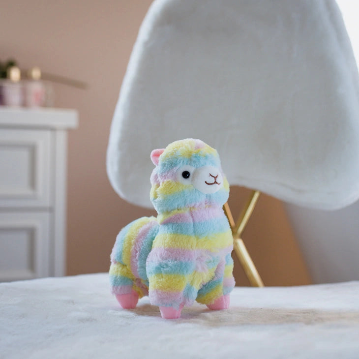 The ZoDoll Rainbow Alpaca Plush Toy, a cute stuffed alpaca plush doll in pastel colors, stands on a white surface in front of a modern white chair and a small cabinet.