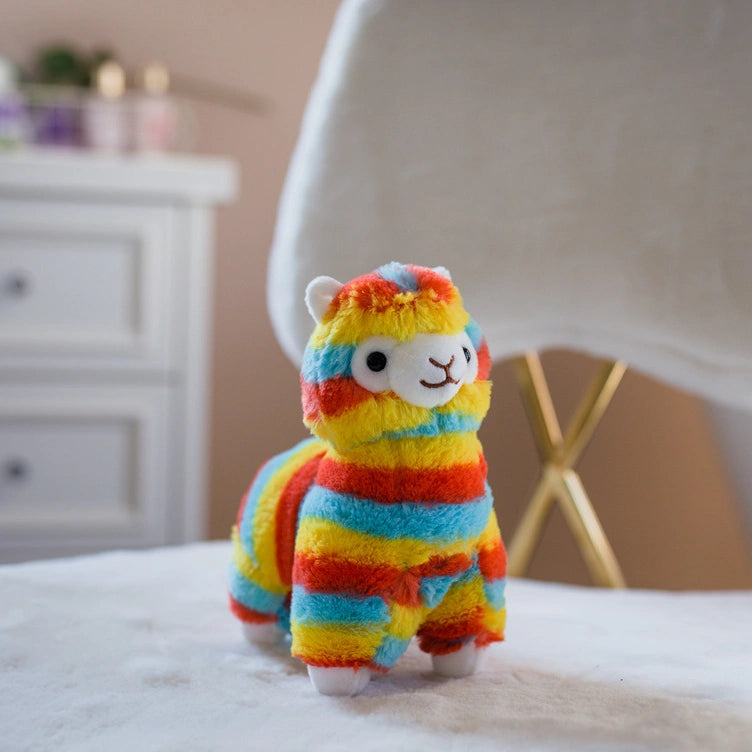 The ZoDoll Rainbow Alpaca Plush Toy, a cute stuffed alpaca plush doll with colorful rainbow stripes, stands on a white furry surface in front of a white chair and drawer.