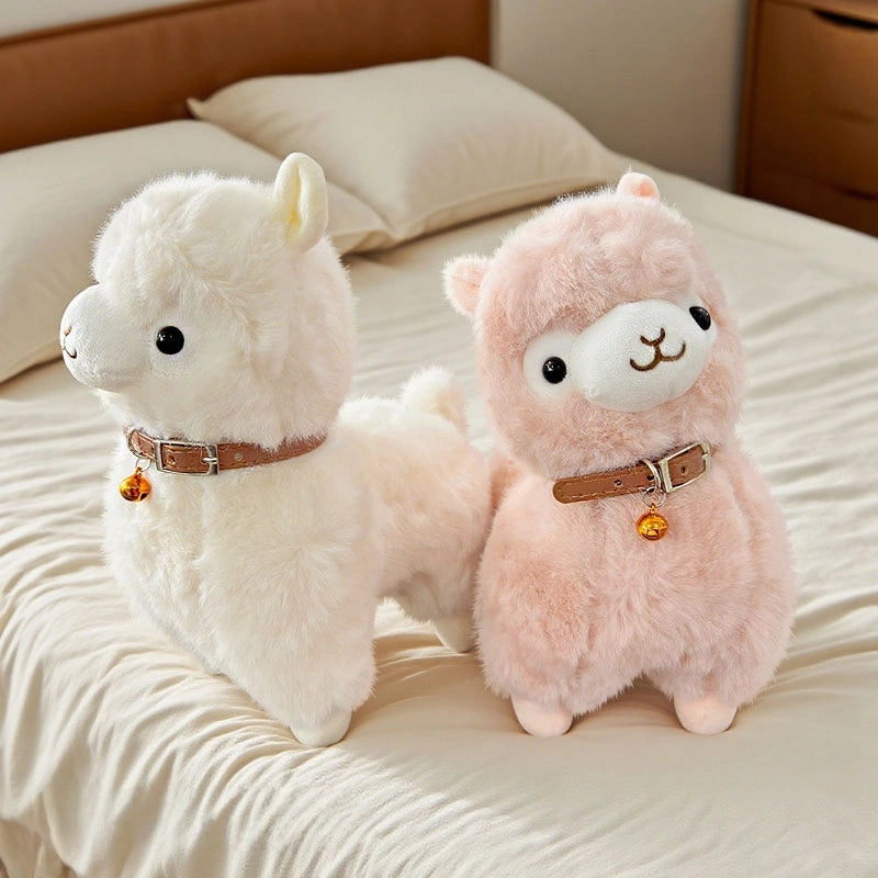 Two ZoDoll Cute Alpaca Plush Dolls—one white, one pink—each with a brown collar and golden bell, are displayed on a neatly made bed.