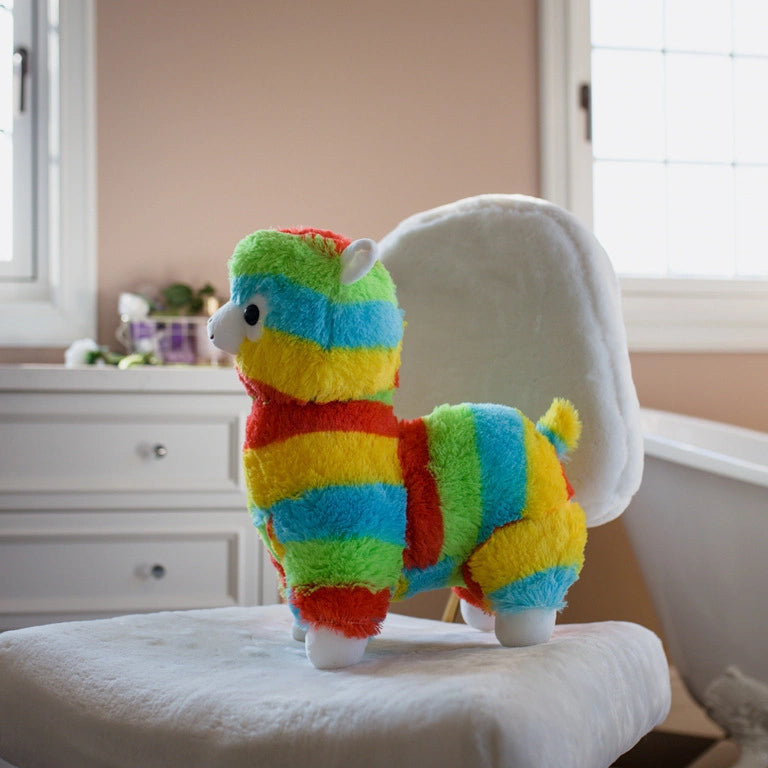 The ZoDoll Rainbow Alpaca Plush Toy – a cute stuffed alpaca doll with colorful stripes – sits on a white chair in a softly lit room with two windows and white furniture.