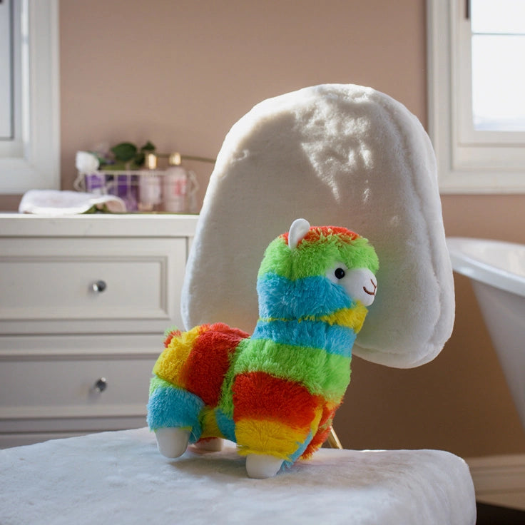 The ZoDoll Rainbow Alpaca Plush Toy – Cute Stuffed Alpaca Plush Doll sits on a white chair in the bathroom, with a window, vanity, and bathtub in the background.