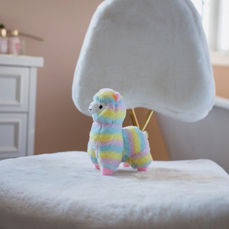 The ZoDoll Rainbow Alpaca Plush Toy—a cute stuffed alpaca doll—sits on a white, fuzzy chair in a softly lit room, its rainbow colors adding a cheerful touch.