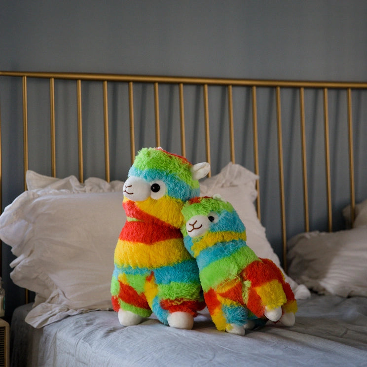 Two ZoDoll Rainbow Alpaca Plush Toys, including a cute stuffed alpaca plush doll, sit on a neatly made bed with white pillows and a brass headboard.