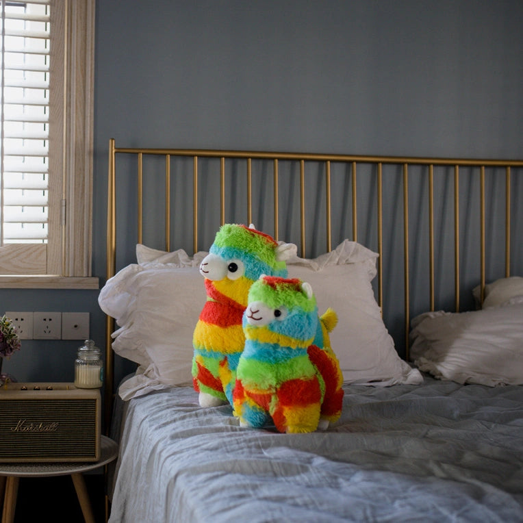 Two ZoDoll Rainbow Alpaca Plush Toy - Cute Stuffed Alpaca Plush Dolls are placed side by side on a neatly made gray bed with a gold headboard.