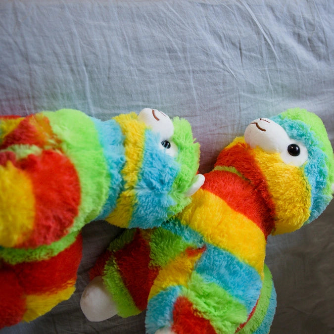 Two ZoDoll Rainbow Alpaca Plush Toy - Cute Stuffed Alpaca Plush Dolls with vibrant, striped fur sit on a light gray fabric surface, facing each other in adorable kawaii style.