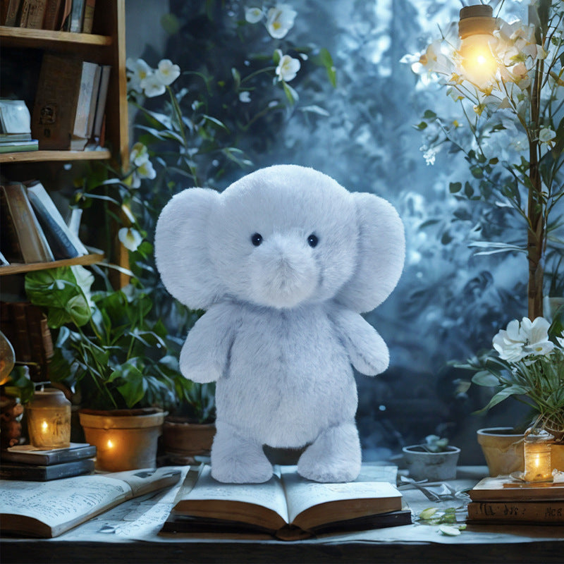 The JOOKI 23cm Blue Elephant Plush Toy, with big ears and a cute face, sits amid open books, potted plants, and candles—a cozy pick for stuffed animal fans or those seeking thoughtful baby gifts.
