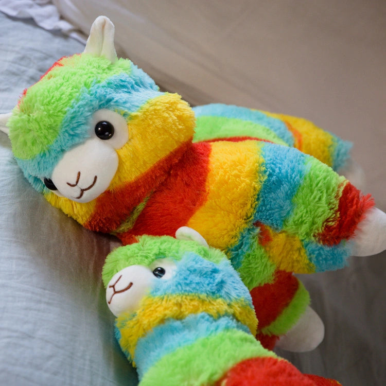 Two ZoDoll Rainbow Alpaca Plush Toy dolls with kawaii features are lying on a bed, while a white pillow is in the background.