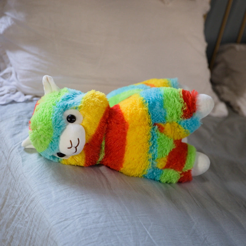 The ZoDoll Rainbow Alpaca Plush Toy, a cute stuffed alpaca plush doll with rainbow-striped fur, is placed on a neatly made bed with white pillows and a light blue sheet.