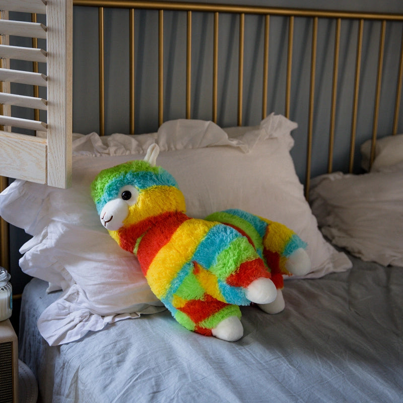 The ZoDoll Rainbow Alpaca Plush Toy, a cute stuffed alpaca plush doll, rests on white pillows atop a neatly made bed with a gray blanket and brass headboard.