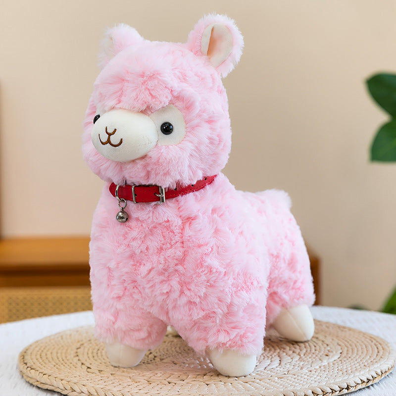 The ZoDoll Cute Alpaca Plush Toy, a soft stuffed alpaca plush with a red collar and bell, is displayed on a woven mat in front of green leaves and a neutral indoor background.