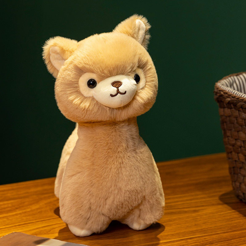 The ZoDoll Cute Alpaca Plush Doll, featuring a soft light brown body and white face, stands on a wooden surface next to a woven basket.