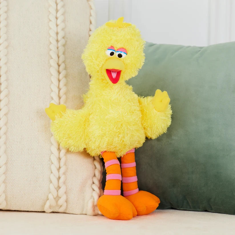A ZoDoll Sesame Street Big Bird Plush – 14 Inch Big Bird Stuffed Animal for Kids and Collectors, featuring yellow fur and orange legs with pink stripes, sits on a light-colored sofa next to a green pillow.