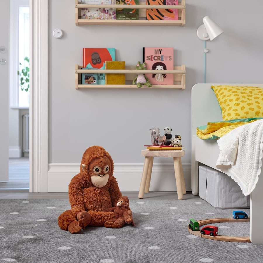 A children’s bedroom with a ZoDoll Giant Punch Monkey Plush Toy - Viral Djungelskog Orangutan hugging pillow on the grey dotted carpet, wooden wall shelves filled with books, and a bed topped with cheerful yellow bedding.