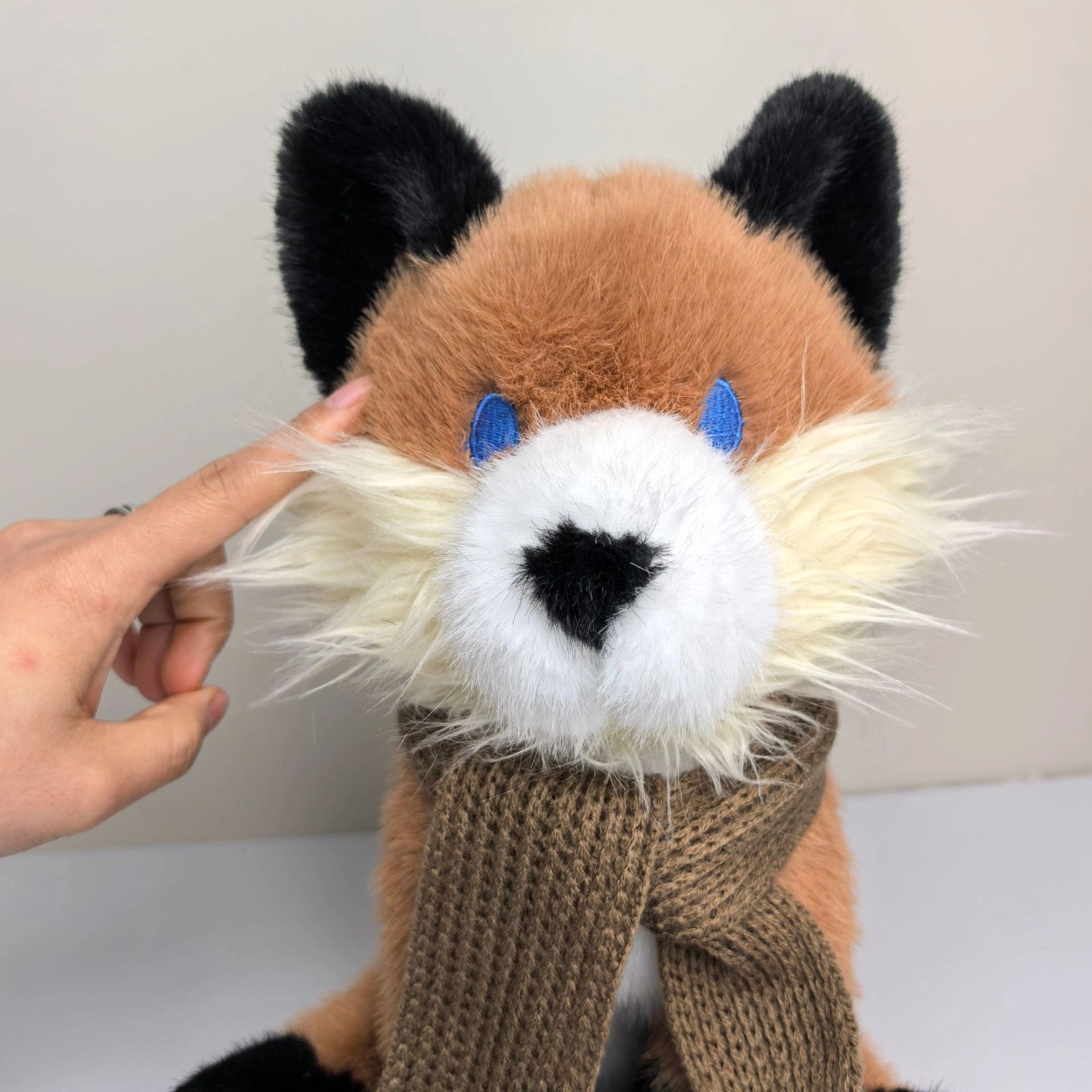 A hand touches the cheek of the ZoDoll Fox Plush Toy, a 30cm soft red stuffed fox with blue eyes, black ears, and a brown scarf—ideal for woodland nursery decor.