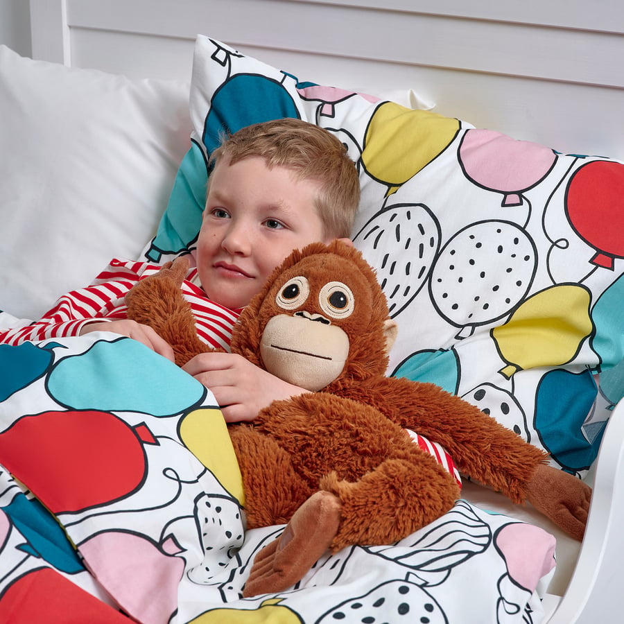 A young child lies in bed on balloon-patterned bedding, cuddling the ZoDoll Giant Punch Monkey Plush Toy—a viral Djungelskog orangutan stuffed animal.