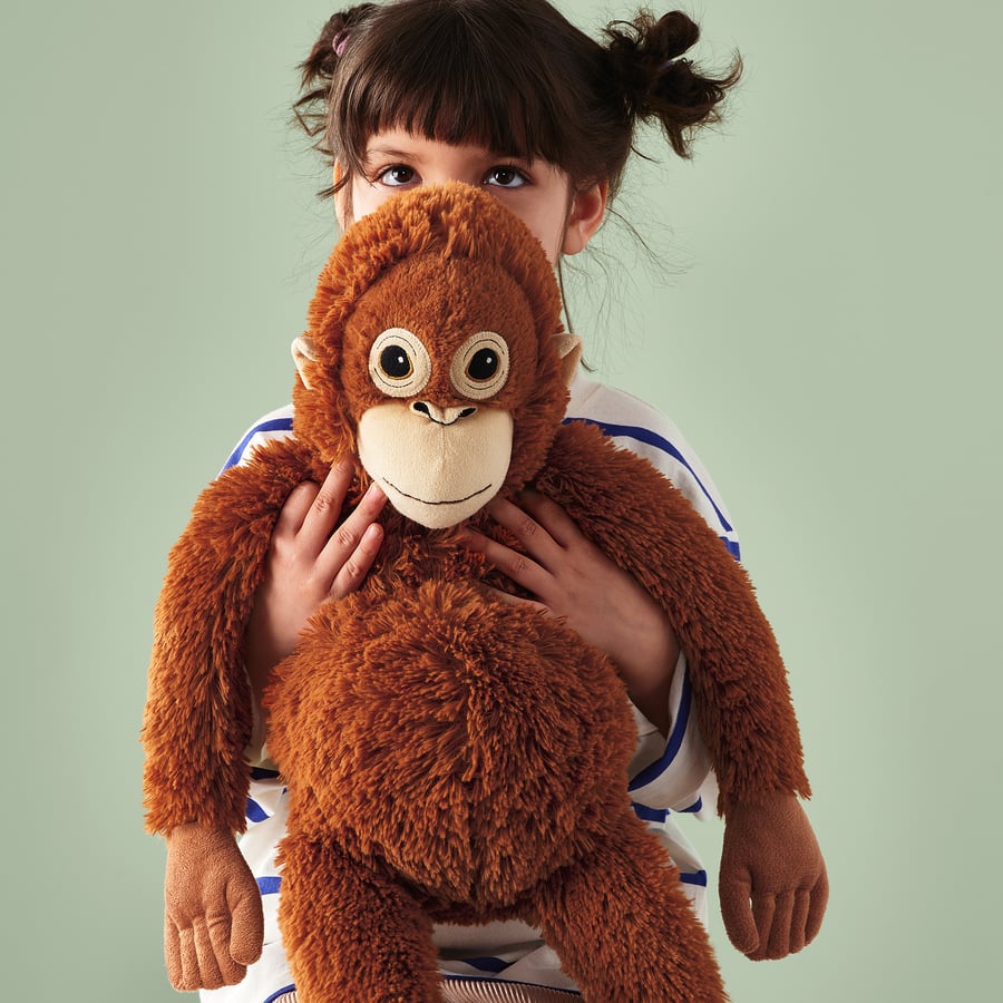 A young girl with brown hair hugs the ZoDoll Giant Punch Monkey Plush Toy—Viral Djungelskog Orangutan Stuffed Animal—close to her chest against a plain light green background.