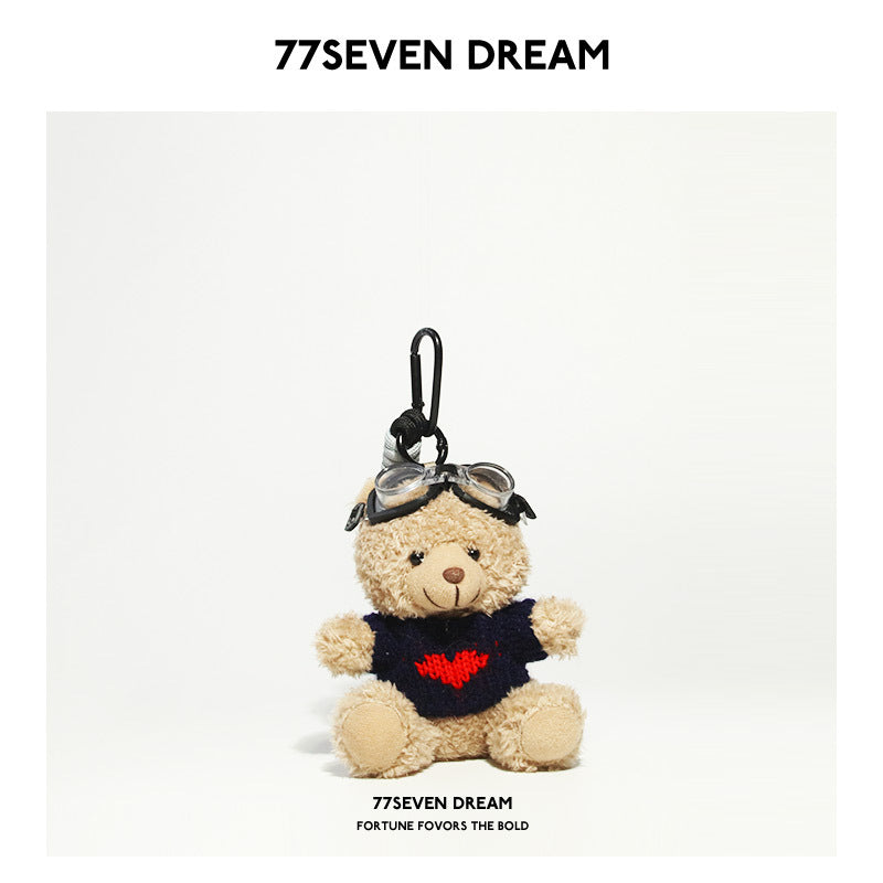 Beige teddy bear plush keychain in a dark navy knit shirt with a red heart graphic and aviator goggles