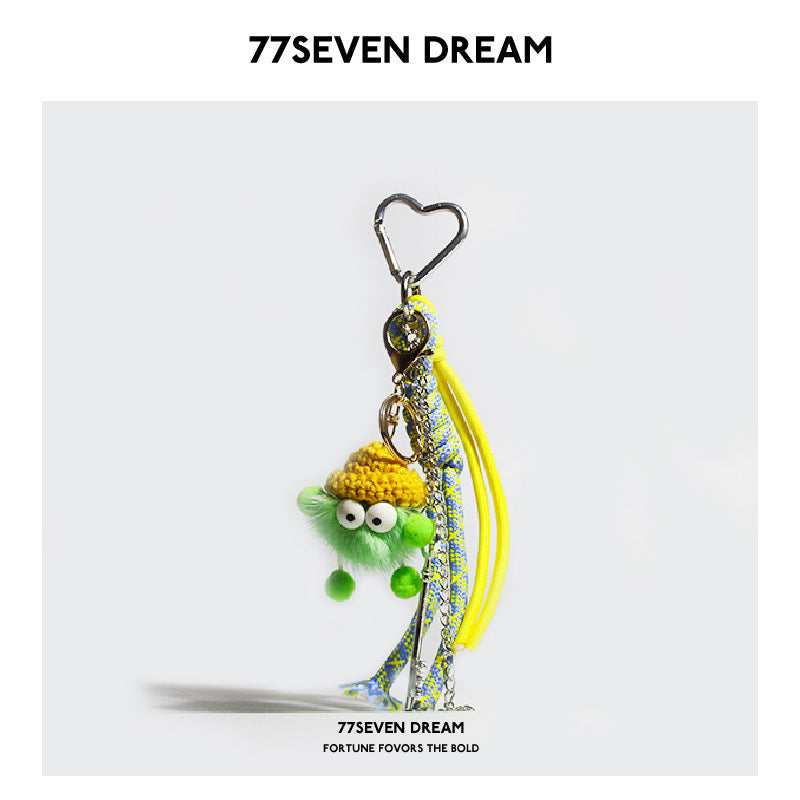 Green fluffy plush monster keychain charm with yellow accent hat, heart carabiner, and multicolored cords