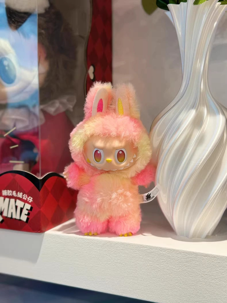 The POP MART Labubu “Loyalty” Plush Keychain from The Monsters “Big Into Energy” Series, a tie-dye bunny bag charm, stands next to a white decorative vase on a shelf.