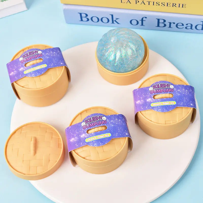 Four round beige ZoDoll Glitter Squishy Dumpling Sensory Fidget Toy containers with steamer are on a white tray; one is open to reveal a blue glittery squishy toy inside.