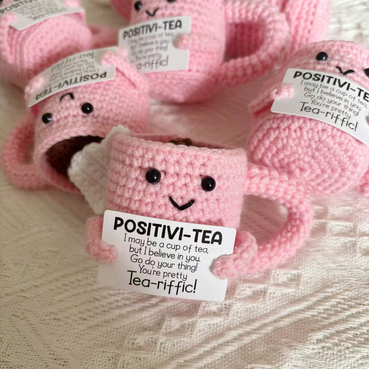 Four ZoDoll Positivi-Tea Crochet Mugs, each with a smiling face and uplifting tea-themed sign, sit on a textured white surface—a delightful amigurumi gift for any tea lover seeking daily encouragement.