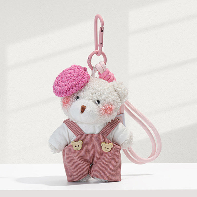 White curly plush bear charm wearing pink corduroy overalls and a pink knitted beret, with carabiner clip and wrist strap