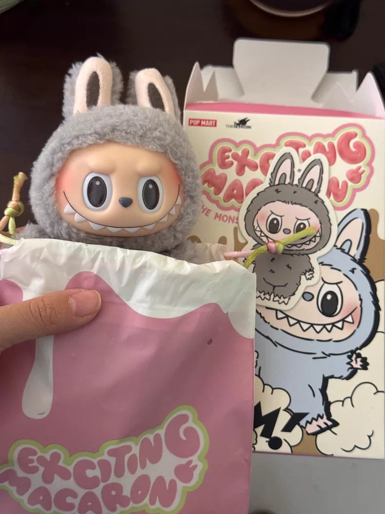 A hand holds a POP MART Labubu “Sesame Bean” Plush Keychain – Standing Grey Bunny Collectible by Kasing Lung, partly in a pink bag, with its matching character art packaging visible in the background.