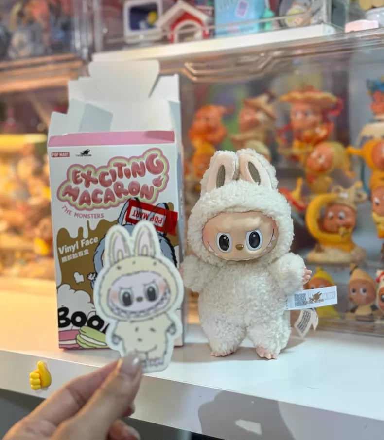 A small POP MART The Monsters Beige Bunny Labubu doll stands beside a white bunny sticker and Exciting Macaron box, with collectible figures and the Labubu "Soymilk" Plush Keychain displayed in the background.
