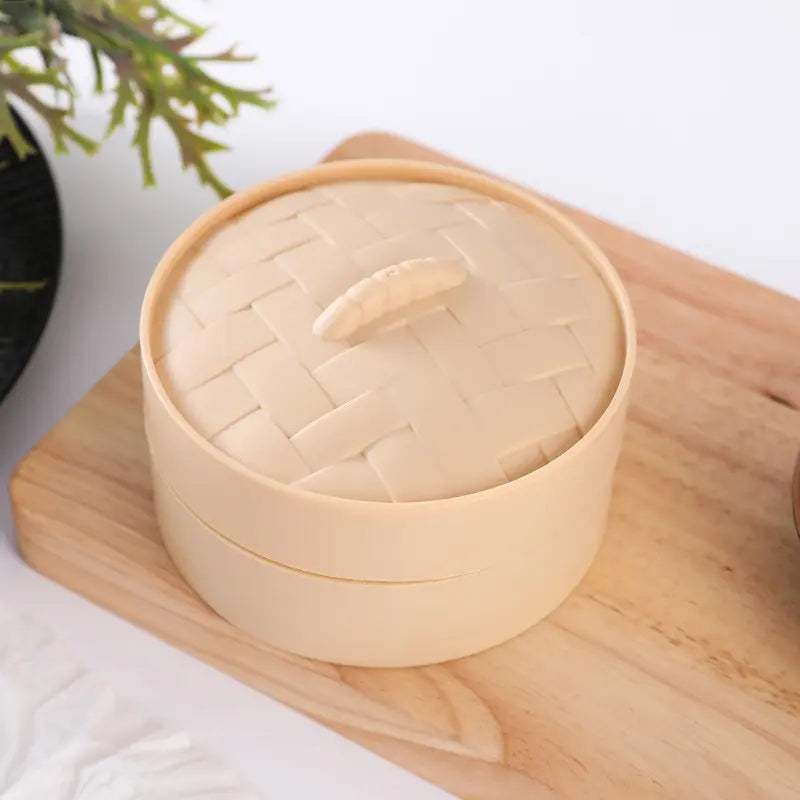 A ZoDoll Giant Emoji Squishy Dumpling Bun Sensory Fidget Toy sits in a bamboo steamer on a wooden board, with a plant and napkin nearby—perfect for stress relief and fun for both kids and adults!.