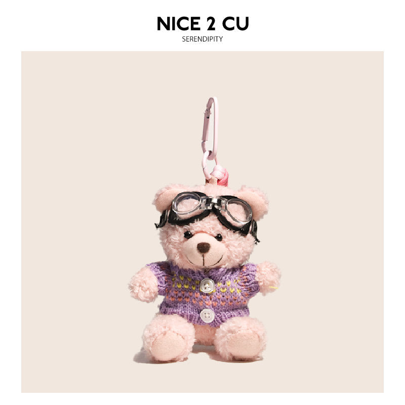 Pink teddy bear plush keychain wearing a purple knit sweater with multicolor accents and black goggles