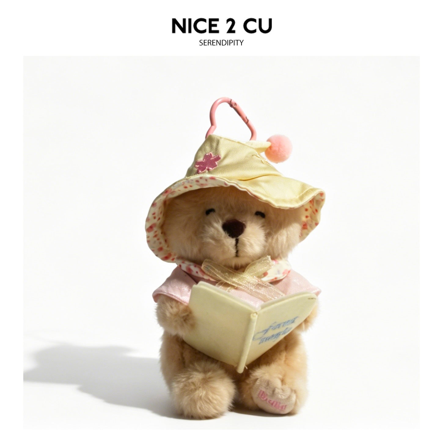 Plush teddy bear charm holding an open book, wearing a hat and outfit, with pink heart-shaped clip