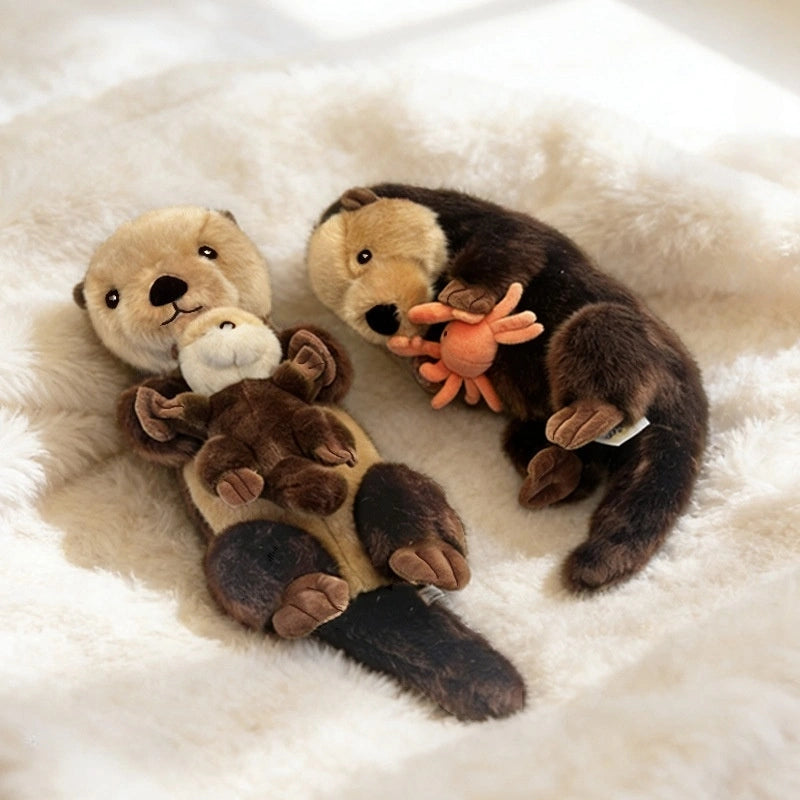Two ZoDoll Otter Plush Toy Soft Sea Otter Stuffed Animals (40x12cm) rest on a soft white blanket; one hugs a removable baby, and the other cuddles a removable orange crab.