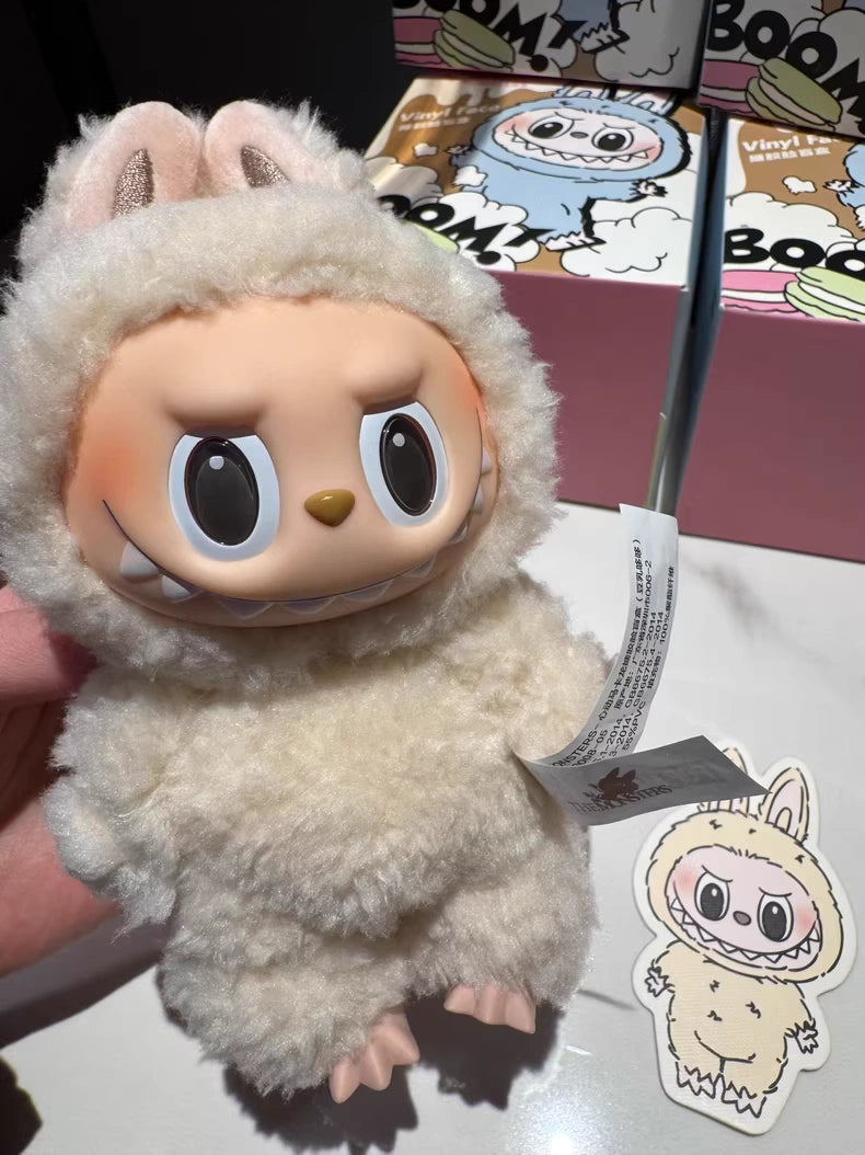 The Labubu “Soymilk” Plush Keychain – Pop Mart The Monsters Beige Bunny Doll by POP MART, with big eyes and animal ears in a fluffy white suit, is held in hand. A matching sticker and colorful character box are in the background.
