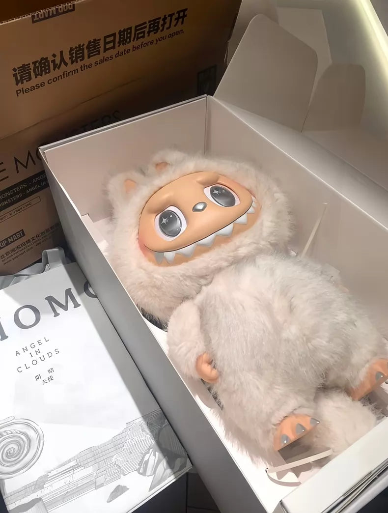 The POP MART Labubu Angel in Clouds 58cm Designer Plush Doll – THE MONSTERS Premium Limited Edition, featuring big eyes and a furry white costume, is displayed in an open box beside a MOMO book and its cardboard packaging.
