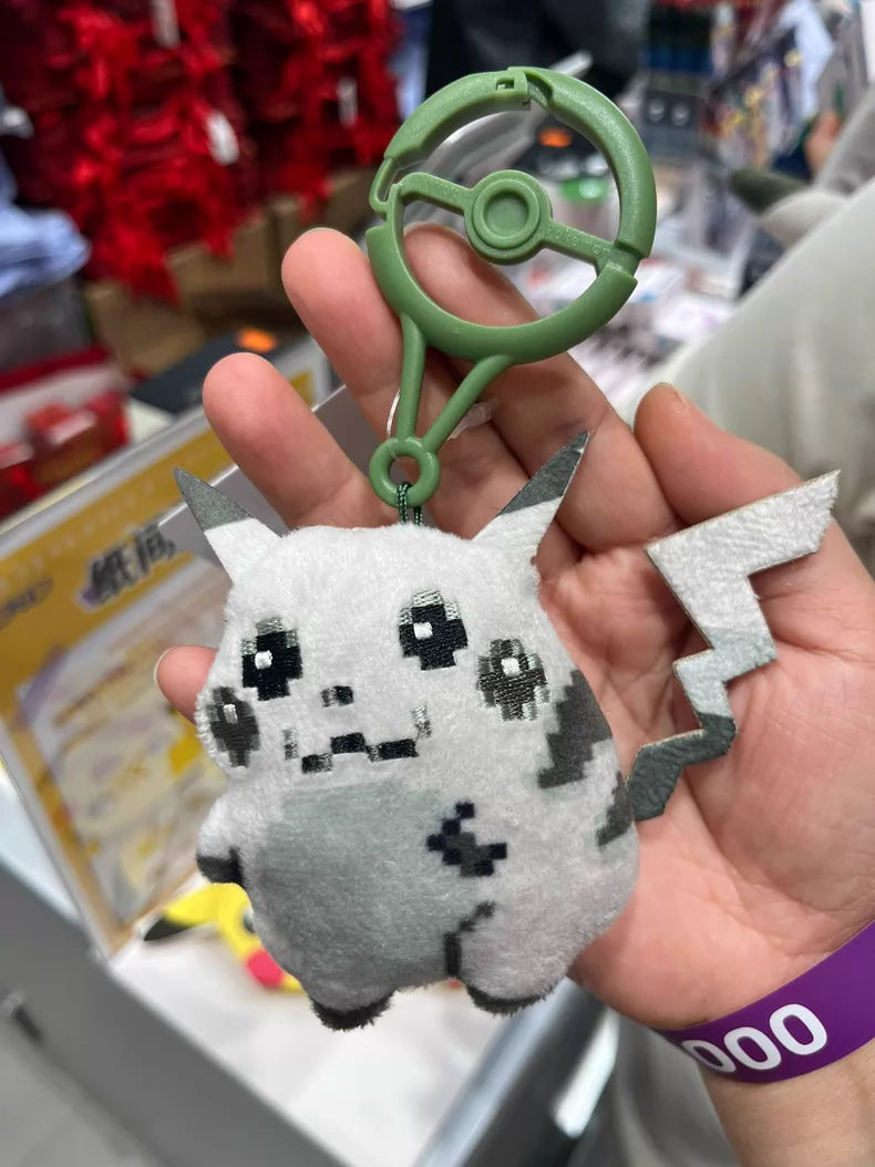 A hand holds the ZoDoll Retro 8-Bit Pikachu of Beginnings Plush Bag Clip (12cm), a classic chunky Pokémon 30th Anniversary keychain, with a green Poké Ball clip—ideal for fans and collectors. Store shelves and merchandise fill the background.