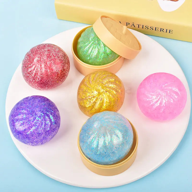 Six glittery, round ZoDoll Glitter Squishy Dumpling Sensory Fidget Toys are displayed on a white plate—two in beige steamers. A yellow Pâtisserie box is visible in the background.