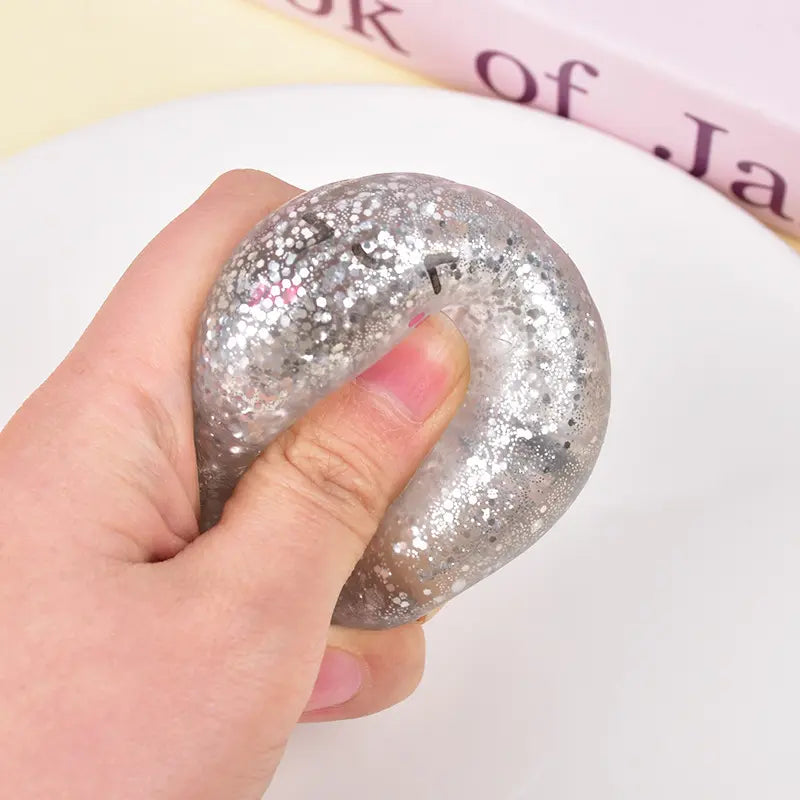 A hand squeezing the ZoDoll Glitter Squishy Dumpling Sensory Fidget Toy with Steamer on a white surface, with a glimpse of a book in the background.