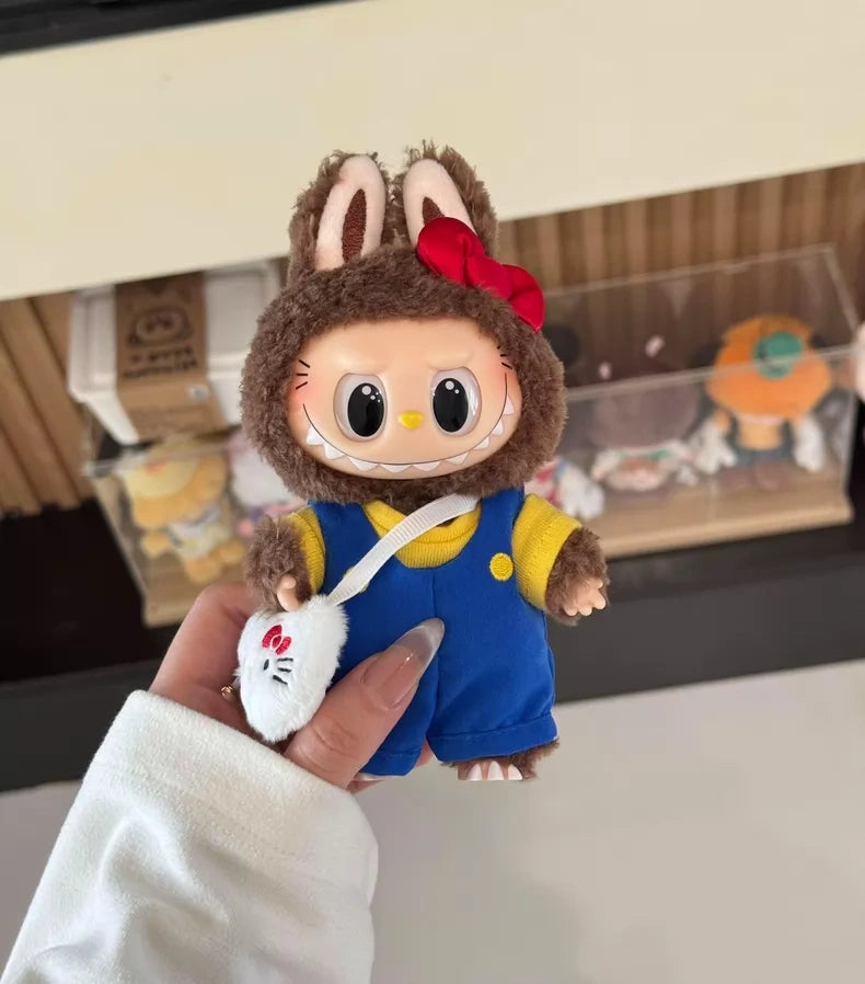 A hand holds the POP MART LABUBU Hello Kitty and Friends Series Secret Edition Plush Pendant Figure—a collectible featuring a small doll in a brown bunny suit with a red bow, blue overalls, and a white pouch with red design.