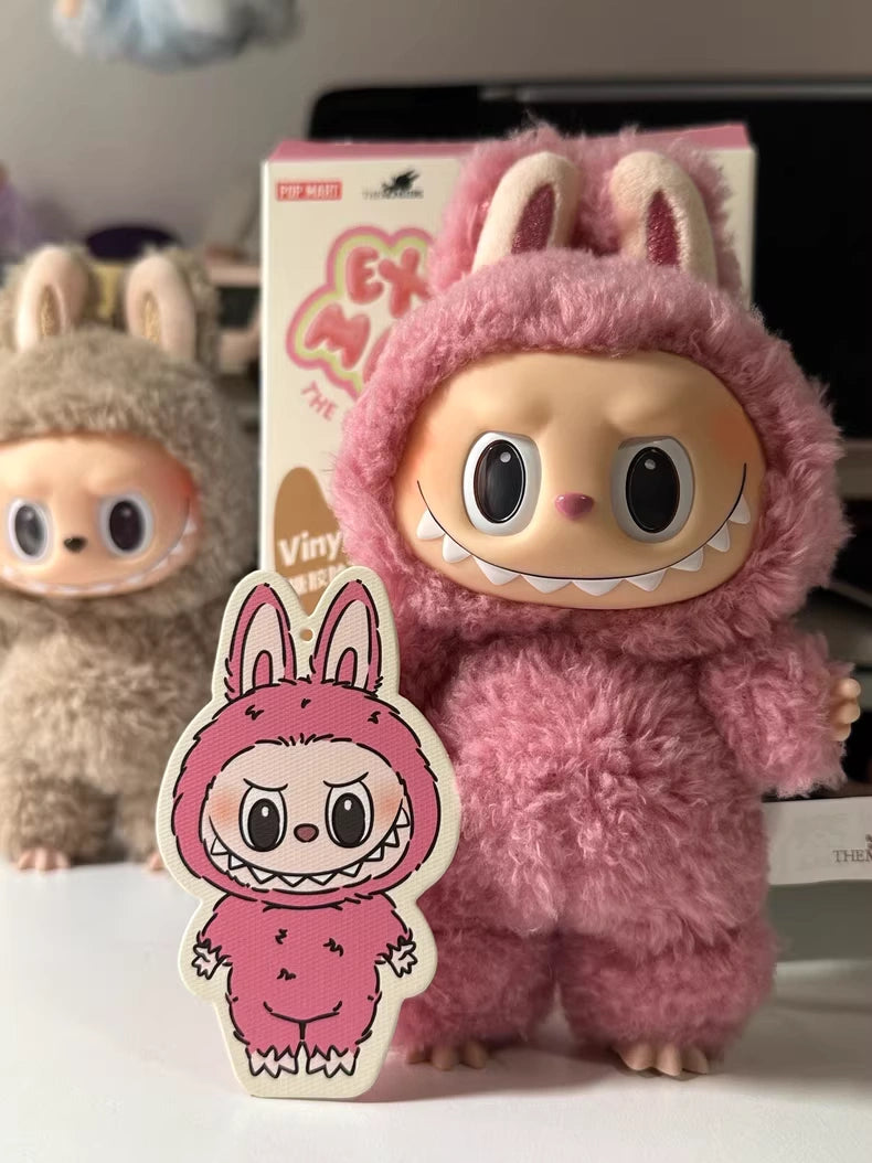 Two POP MART Labubu “Lychee Berry” Plush Keychain dolls in pink fleece bunny costumes stand on a table. The pink Pop Mart The Monsters doll holds a matching cartoon cutout, while another plush stands further back.