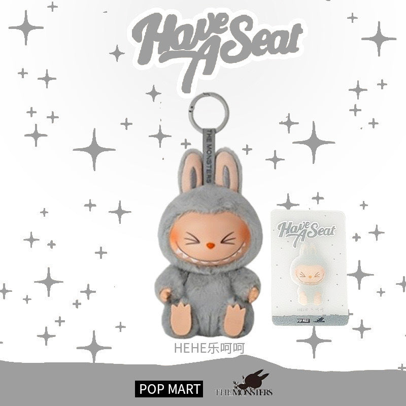 The Labubu “Hehe” Plush Keychain – Have a Seat Series by POP MART features The Monsters’ character in a gray bunny suit with closed eyes, displayed with packaging and "Have A Seat" text above against a starry background.